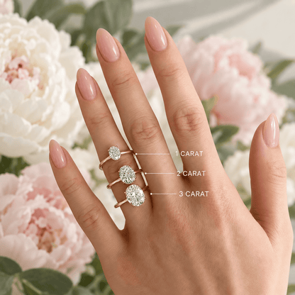 A close-up of a hand wearing three gold plated rings with oval-cut moissanite of varying sizes, labeled '1 CARAT', '2 CARAT', and '3 CARAT' 