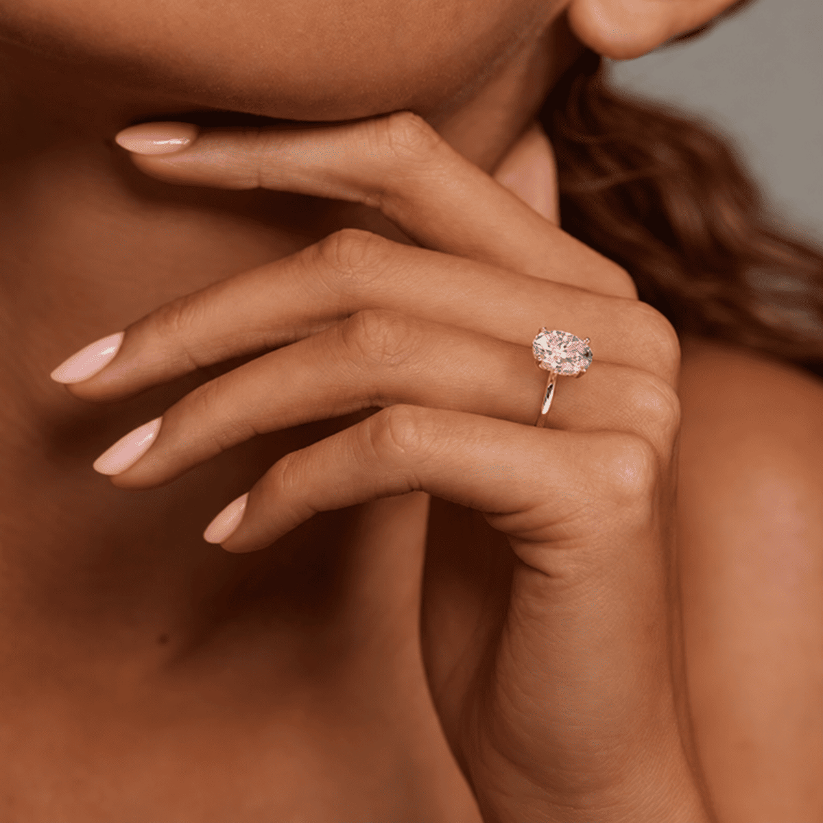 ANNXMEE Celeste oval moissanite engagement ring worn on a hand beneath the chin, with a pale peach-pink manicure.