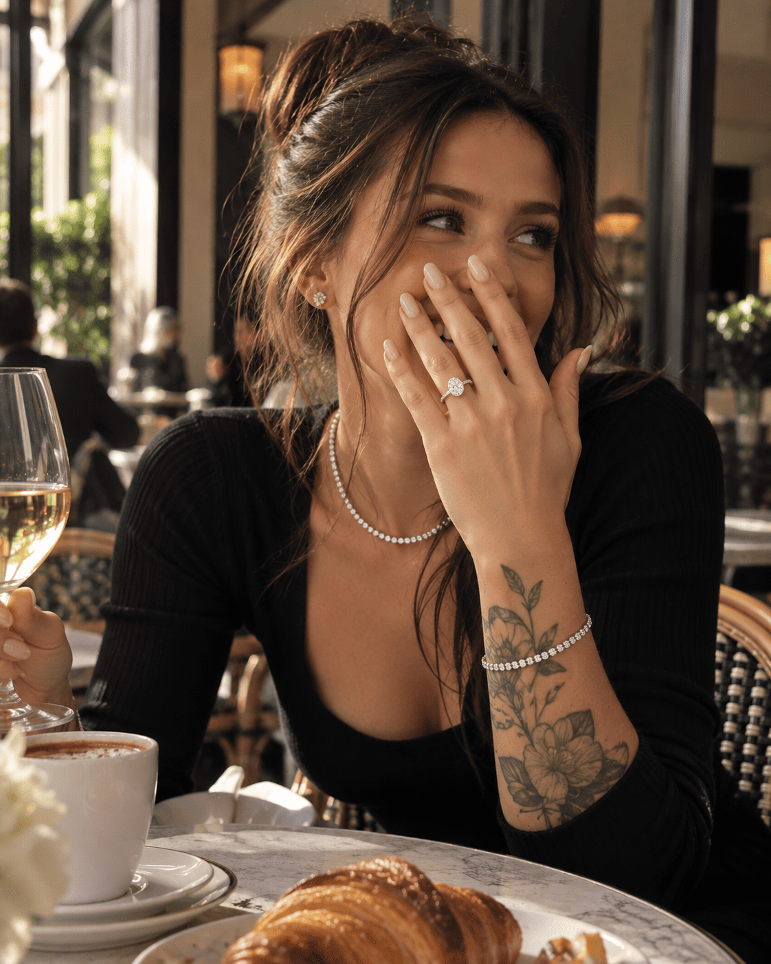 A woman in a black ribbed top sits at a cafe table, smiling and covering her mouth while wearing a diamond ring on her finger, a pearl necklace, and a pearl bracelet. A croissant and coffee cup are on the table in front of her. ANNXMEE Moissanite Ring
