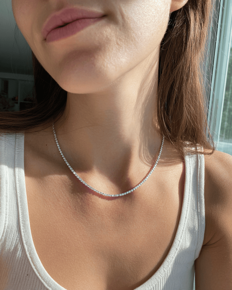 A close-up lifestyle shot of a woman wearing a delicate silver tennis necklace with small clear stones, paired with a white ribbed tank top. ANNXMEE Moissanite Tennis Necklace 