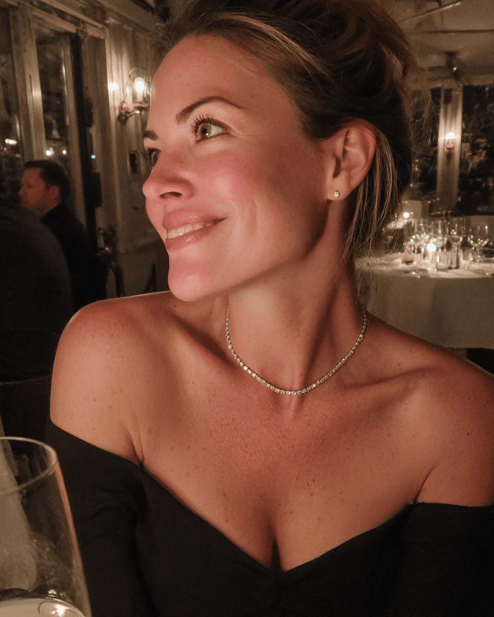 A woman in a black off-the-shoulder dress wearing a delicate silver beaded necklace and a small stud earring, photographed in a warmly lit restaurant setting. Moissanite Earrings. ANNXMEE