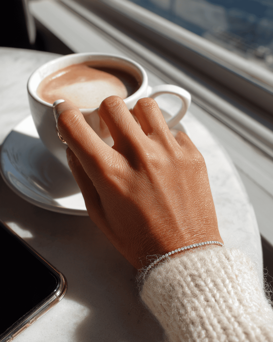 A close-up lifestyle shot of a person's hand resting on a white saucer next to a cup of coffee, wearing a delicate silver chain bracelet on the wrist. ANNXMEE Moissanite Tennis Bracelet