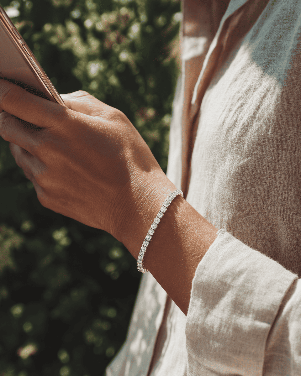 A close-up lifestyle shot of a woman wearing a delicate silver tennis bracelet on her wrist, holding a smartphone outdoors with a blurred green background. Moissanite Tennis Bracelet. ANNXMEE Jewelry 